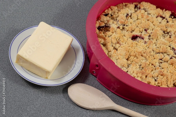 Fototapeta Frischer Pflaumenkuchen in Silikonbackform neben einem Stück Butter und einem Kochlöffel aus Holz
