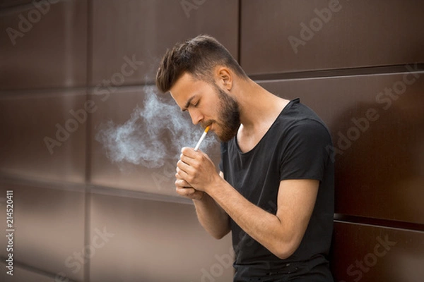 Fototapeta A young handsome white bearded man in black t-shirt smokes a cigarette in the street in the spring. Close up.