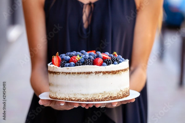 Obraz The girl is holding a cake with forest berries, strawberries, blueberries, on the street in the city. A concept for the production of cakes, bakeries, restaurants. Background image