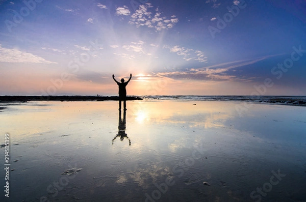 Fototapeta Silhoutte of man raise hand above facing the sunrise with water reflection
