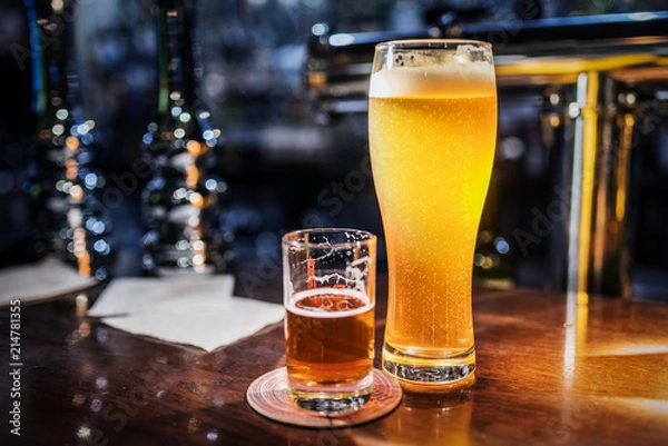 Obraz Light beer in a beer glass and dark beer in a small glass lighted with warm light on a bar counter