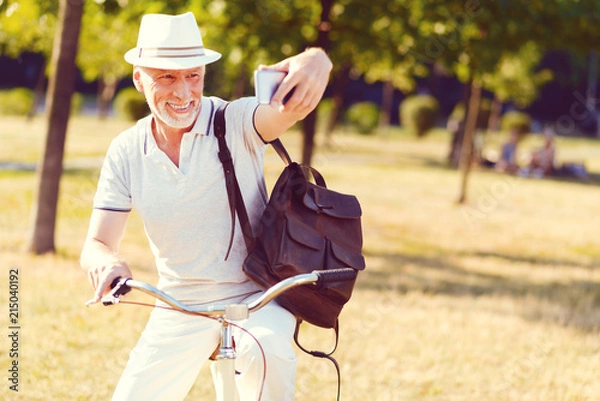 Fototapeta One more shot. Radiant elderly man grinning broadly while taking a break after cycling and taking a selfie in a park.
