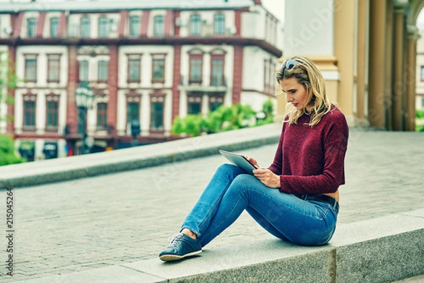 Fototapeta A woman in the city with a tablet .The girl uses new technologies for work and rest