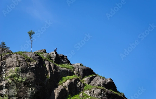 Fototapeta Man Sitting Alone on the Rocks Cliff with Isolated Tree Blue Sky in Summer Scotland UK