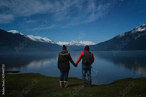 Fototapeta Couple holding hands standing on lake looking the night starry sky and mountain reflection in Patagonia Argentina