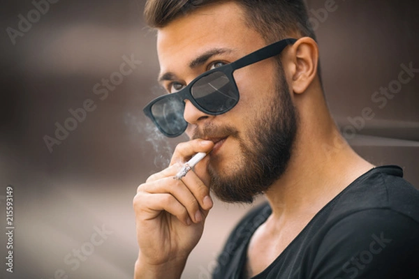 Fototapeta A young handsome white bearded man in black t-shirt smokes a cigarette in the street in the spring. Close up.