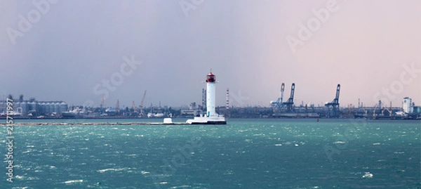 Fototapeta Lighthouse in the sea against the backdrop of a cargo port. Summer seascape with a white lighthouse with red top. Black Sea. Sea Port of Odessa.