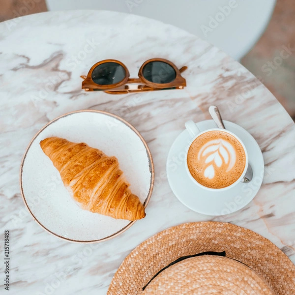 Fototapeta Morning cup of coffee with beautiful latte art and crossant, straw boater hat and brown sunglasses on the round marble table. Flatlay, istagram style.
