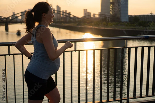 Obraz Active pregnant woman running across the bridge with a beautiful view of the city of Frankfurt in Germany