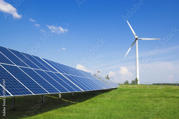 Fototapeta wind generator of electricity from three blades and solar panels of a battery of photocells against a background of clouds and a blue sky green grass