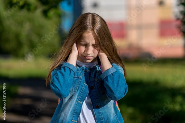 Fototapeta Little girl schoolgirl in summer park. He covers his ears with hands. The concept of loud noise, problems in family, scandal, reluctance listen, discontent. Emotions of quarrel, frustration, conflict.