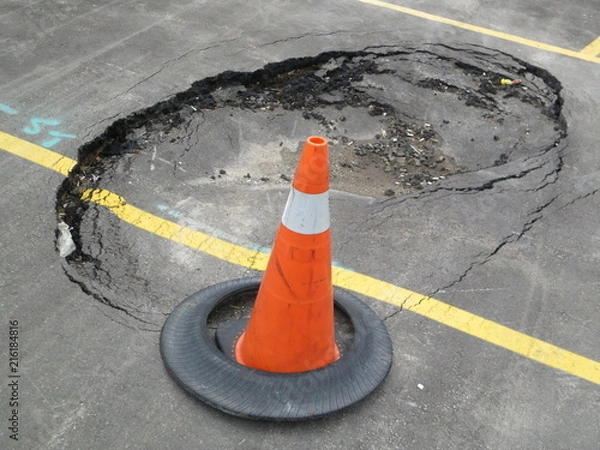 Obraz Constructions at unexpected sink hole in public parking lot with traffic cone. This accident causes damage or injuries. It needs instant attention and repair work and proper foundation of the ground.