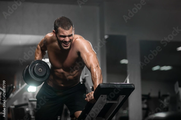 Fototapeta Man trains in the gym. Athletic man trains with dumbbells, pumping his biceps
