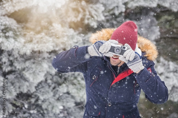 Fototapeta Fashionable man taking pictures with retro camera on snowy day. Winter vacation travel concept.