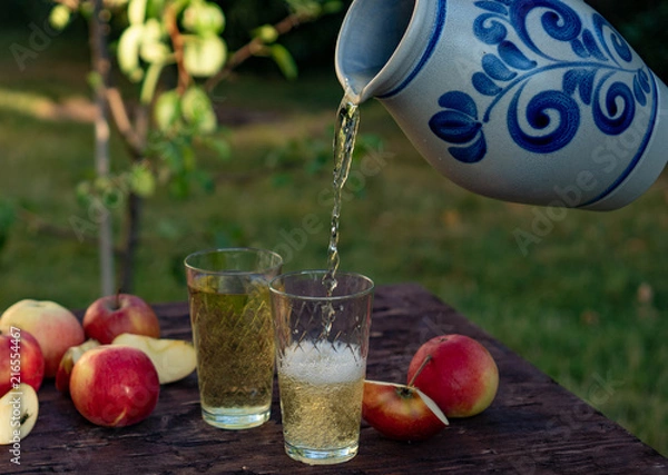 Obraz a man's hand pours Traditional apple wine in a refilled glass in the city of Frankfurt. A jug of wine on an old wooden table in the garden, around it apples
