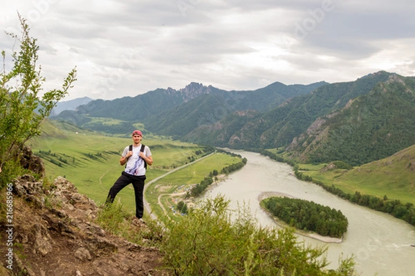 Fototapeta A young guy in a cap with a backpack and black sports pants is standing on the edge of a cliff in the mountains of the Altai, from where he opens a view of the river Katun and the island