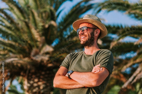 Fototapeta Young man standing by the palm trees looking at sun