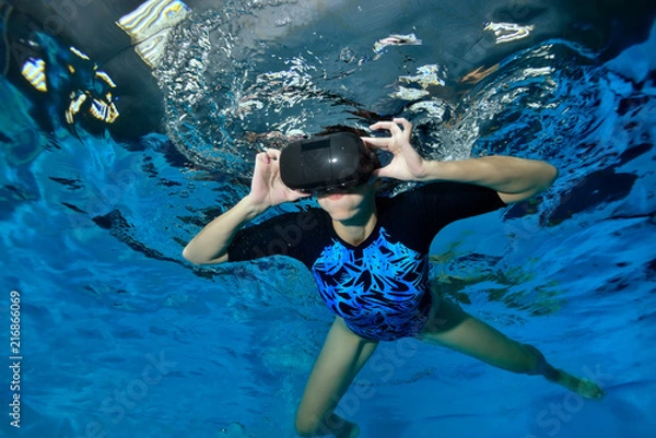 Obraz Virtual reality simulator. An unusual girl swims underwater in the pool in black virtual reality glasses on her head. Shooting underwater. Horizontal orientation