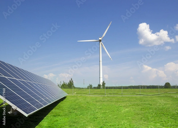 Fototapeta wind generator of electricity from three blades and solar panels of a battery of photocells against a background of clouds and a blue sky green grass