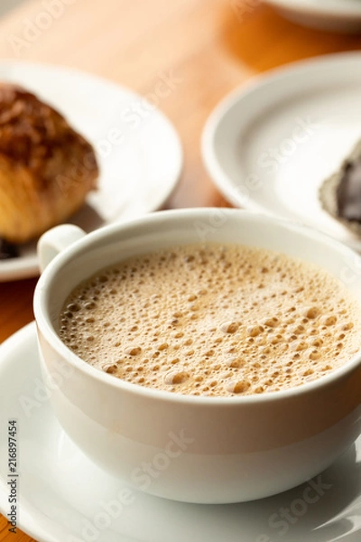 Fototapeta Close up on coffee in a white cup and saucer,  with pastry in the background