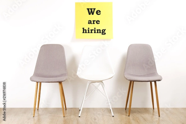 Fototapeta Job recruitment advertisement, WE ARE HIRING text written on yellow wall poster, three empty loft style chairs. Human resources campaign to find workers for vacant job. Close up, copy space background