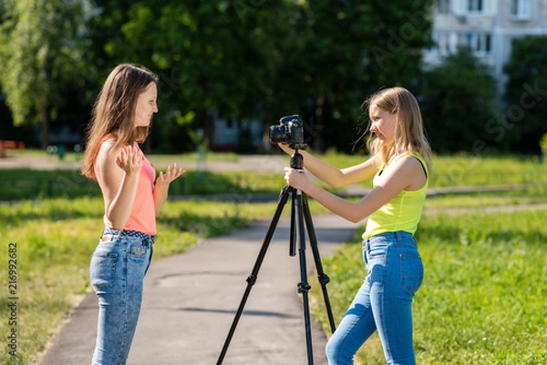 Fototapeta Where little teenage girls in the summer in park are recording video on the camera. Emotionally gesticulates with his hand using a camera with a tripod.