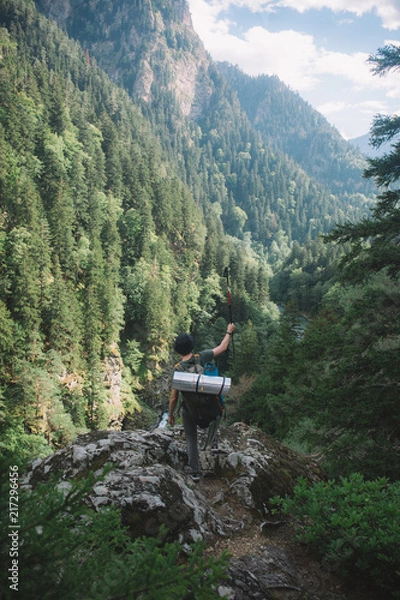 Fototapeta  a young guy with a tourist backpack looks down from the mountain to the river and a thick green forest