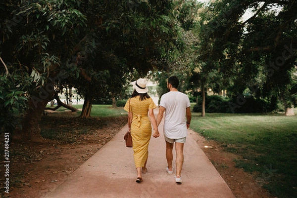 Obraz Stylish cute brunette girl with her boyfriend with beard walking holding hands close to each other on the small road in the park between large trees in Spain in the evening. Date of the young couple