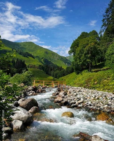 Obraz A mountain river with a bridge with green mountains and blue sky in the background