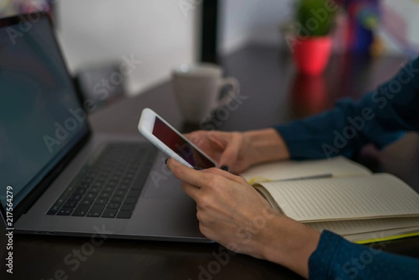 Fototapeta Selective focus on cellphone in hands. Close up view of woman internet user checks notification on mobile phone sitting front laptop computer,female person using modern devices and connecting to netwo