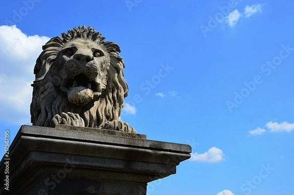Obraz Lying stone lion statue guarding Budapest Chain Bridge, alson known as Szechenyi Landhid, blue summer skies with some clouds in background