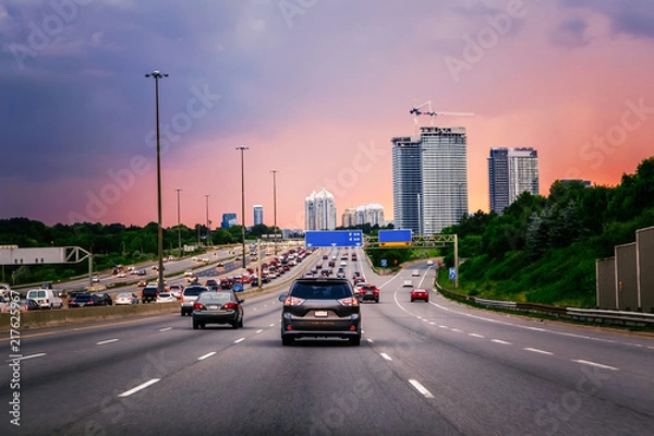 Fototapeta Ruch nocny. Samochody na autostradzie o zachodzie słońca wieczorem w typowym ruchliwym amerykańskim mieście. Piękny, niesamowity nocny widok miejski z czerwonymi, żółtymi i niebieskimi chmurami nieba. Zachód słońca w centrum miasta.