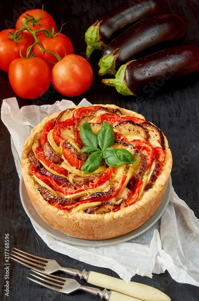 Fototapeta Pie with eggplants, tomatoes and cheese on the gray plate decorated with fresh basil leaves and vintage forks. Homemade vegetable tart on the black background. Side view