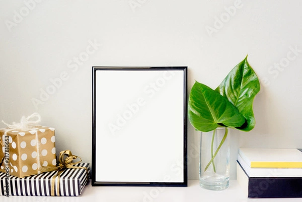 Fototapeta Black photo frame, green plant in a chrystal vase, gift boxes and a pile of books arranged against emty grey wall. Frame mock-up.