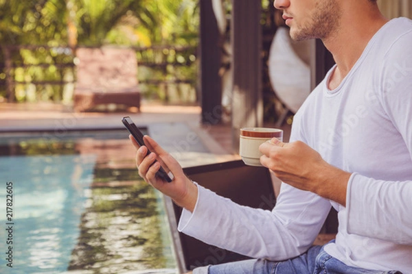 Fototapeta Attractive man using cellphone and drinking coffee while sitting near swimming pool.