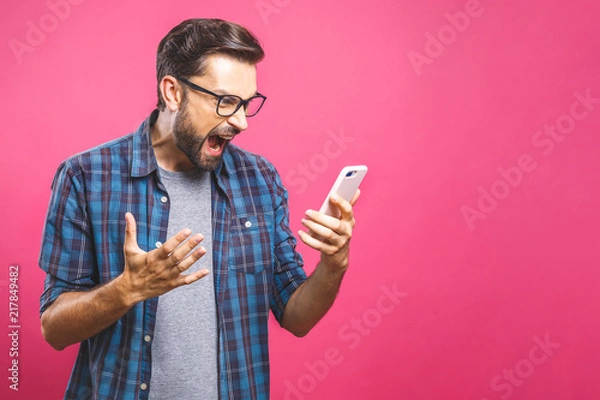 Fototapeta Young caucasian man angry, frustrated and furious with his phone, angry with customer service over pink background.