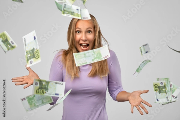 Fototapeta Portrait of cheerful glad girl enjoying shower from 100 euro, flying money, gesturing with hands isolated on gray background