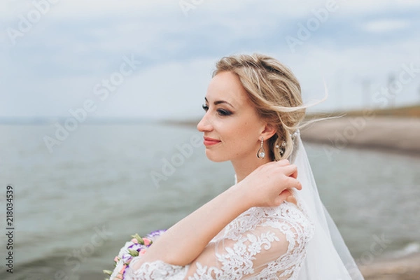 Fototapeta A beautiful bride in a lace dress dreams at the pier, against the background of the sea and sky. Wedding portrait close-up of a curly bride on the waterfront.