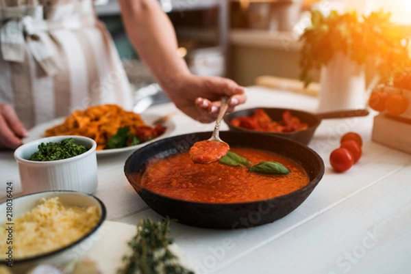Fototapeta Woman making homemade pasta with tomato sous and cheese over old wooden table. Tomato, olive oil, spices, herbs, cheese, tomato sauce on a weathered wooden table in the summer sun's rays. Organic food