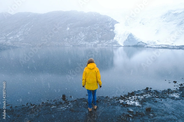 Fototapeta A girl in a yellow jacket and jeans stands on the shore of a blue lake in the snow, on the shore of snowy glaciers