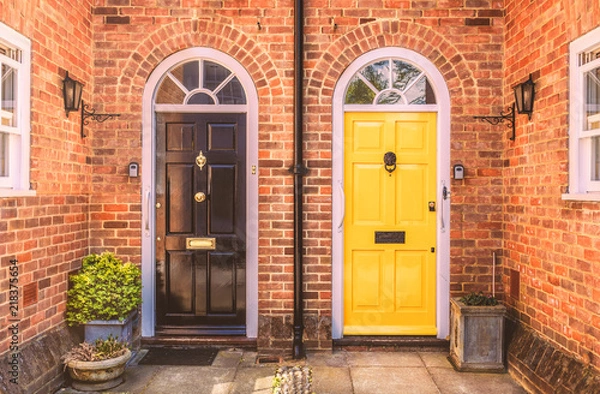 Fototapeta Two residential front doors, one yellow, one black with a drain pipe down the middle. The walls are red brick there are two side windows and lunette arches over the doors