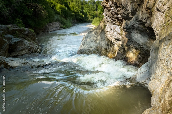 Fototapeta Mountain river with a waterfall flowing between rocky coasts. Over the river trees grow. Taken at sunny spring day at Carpathian mountains. Fast flowing water. Natural background. Freshness feel.