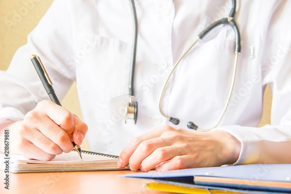 Fototapeta Young female medical student using modern smartphone in the hospital, writing in the notebook. Close up hands shoot
