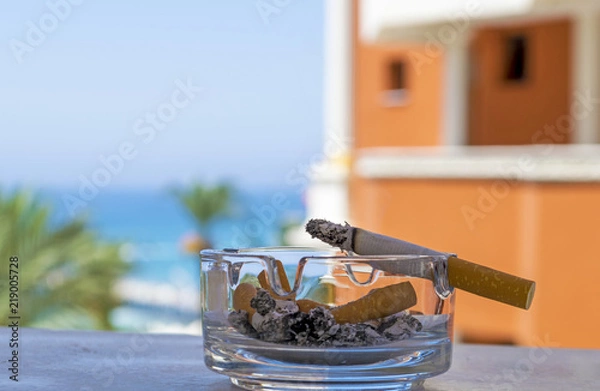 Fototapeta Ashtray with a cigarette on the background of the sea beach. Cigarette lying on a glass ashtray. Cigarette smoldering. 