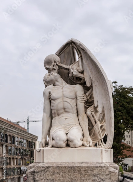 Fototapeta The Kiss of Death statue in Poblenou Cemetery in Barcelona. This marble sculpture depicts death, as a winged skeleton, kissing a handsome young man. The sculpture is at once romantic and horrifying.