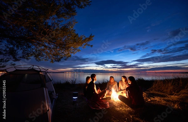 Fototapeta Nocny letni kemping na brzegu morza. Grupa pięciu młodych turystów siedzi na plaży wokół ogniska w pobliżu namiotu pod pięknym niebieskim wieczornym niebem. Turystyka, przyjaźń i piękno koncepcji natury.