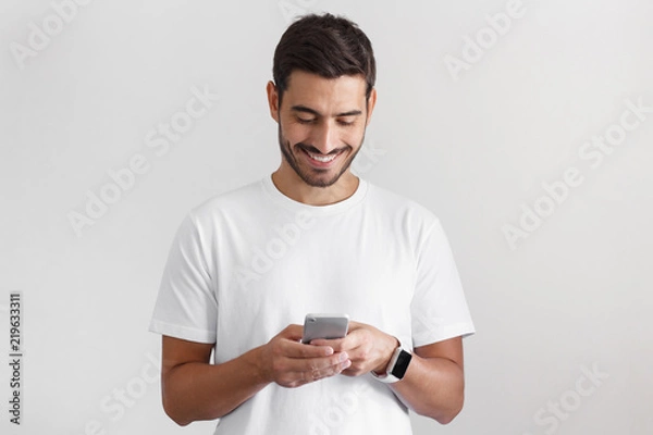 Fototapeta Daylight portrait of young european caucasian man isolated on gray background wearing white t-shirt standing in front of camera, looking attentively with smile at screen of smartphone he is holding