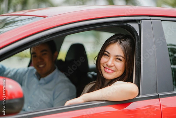 Fototapeta Portrait of a young Japanese Asian woman leaning out the window of a red car window and smiling happily. She is being driven to her destination in a ride she booked on a ride hailing app.