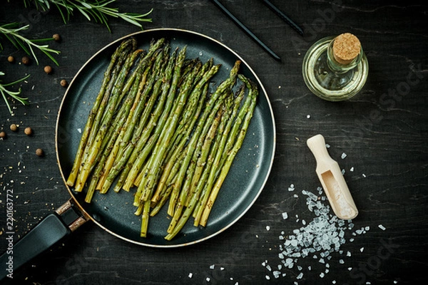 Obraz delicious green asparagus in a pan on a wooden black table with rosemary salt and olive oil