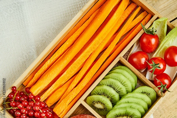 Fototapeta box with delicious carrots, apple, kiwi, cherry tomatoes and redcurrant on a wooden table